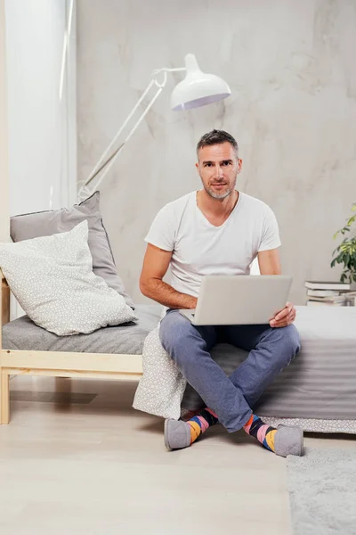 잘생긴 백인 중간 나이 든된 남자 캐주얼 옷을 입고 침실에서 침대에 앉아있는 동안 노트북을 사용 하 여. — 스톡 사진