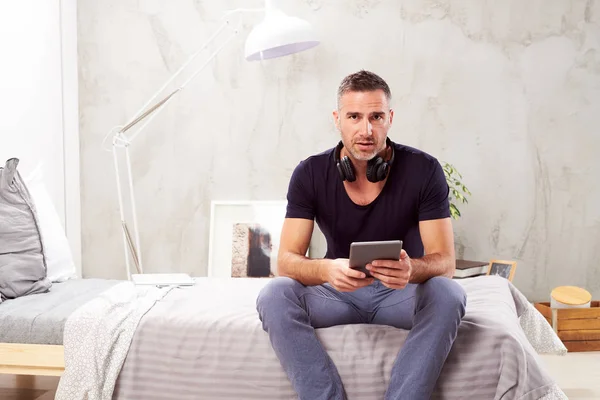 Uomo caucasico sulla quarantina seduto sul letto in camera da letto e utilizzando tablet . — Foto Stock