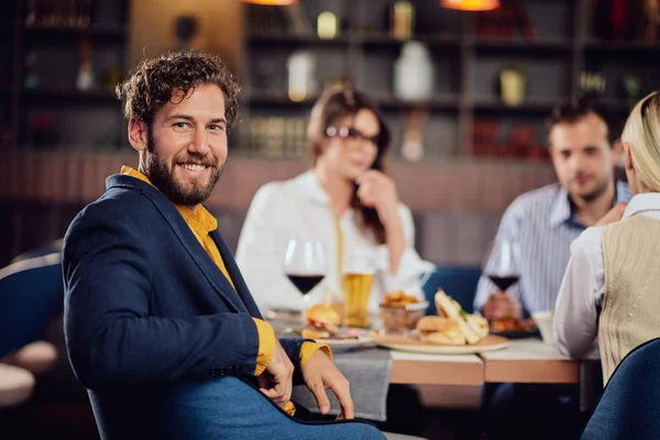 Jonge glimlachende man gekleed slim casual zitten in restaurant en kijken over de schouder. Op de achtergrond zijn vrienden aan het eten. — Stockfoto