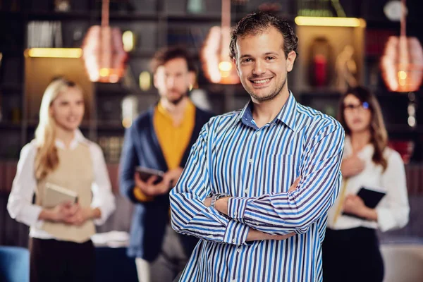 Jonge aantrekkelijke arabische zakenman staat in restaurant met gekruiste armen. Op de achtergrond zijn succesvolle team poseren. — Stockfoto
