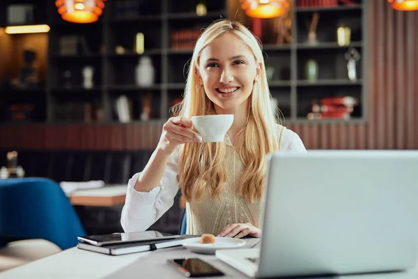 Encantadora mujer de negocios rubia sonriente vestida elegante informal sentada en la cafetería, bebiendo café y utilizando el ordenador portátil . — Foto de Stock