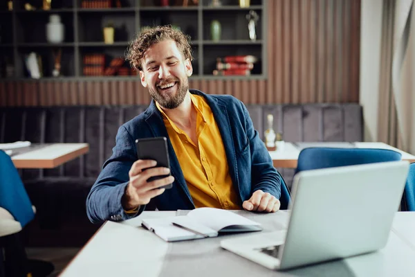 Joven hombre de negocios caucásico vestido casual inteligente usando el teléfono inteligente mientras está sentado en la cafetería. En la computadora portátil de escritorio . — Foto de Stock