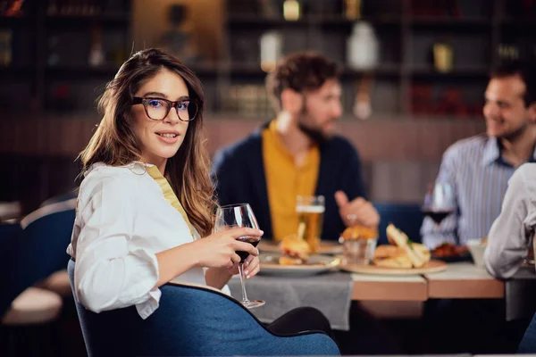 Retrato de linda morena olhando para a câmera e segurando vidro com vinho tinto enquanto está sentado no restaurante. No fundo amigos comendo restaurante . — Fotografia de Stock