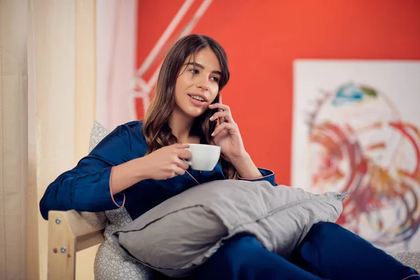 Schöne kaukasische Brünette im Pyjama sitzt auf dem Bett im Schlafzimmer, telefoniert und trinkt ihren Morgenkaffee. — Stockfoto