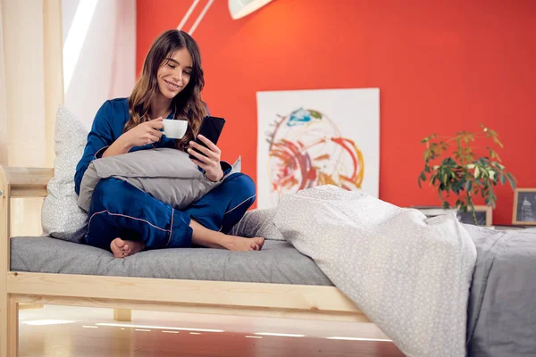 Hermosa morena en pijama azul sentada en la cama en el dormitorio con las piernas cruzadas, bebiendo café de la mañana y usando el teléfono inteligente . — Foto de Stock