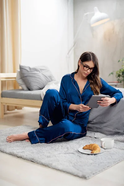 Hermosa morena caucásica en pijama azul y con gafas de vista usando tableta mientras está sentada en el suelo en el dormitorio. En el suelo está Breeakfast. Hora de la mañana . — Foto de Stock