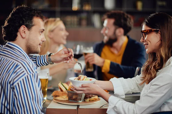 Amigos multiétnicos sentados no restaurante, bebendo álcool, conversando e comendo hambúrgueres para o jantar . — Fotografia de Stock