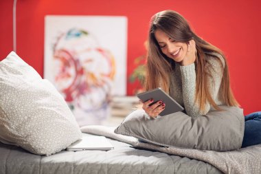 Rahat yatak yatak odasında oturmuş ve tablet kullanma muhteşem gülümseyen beyaz esmer giyinmiş.