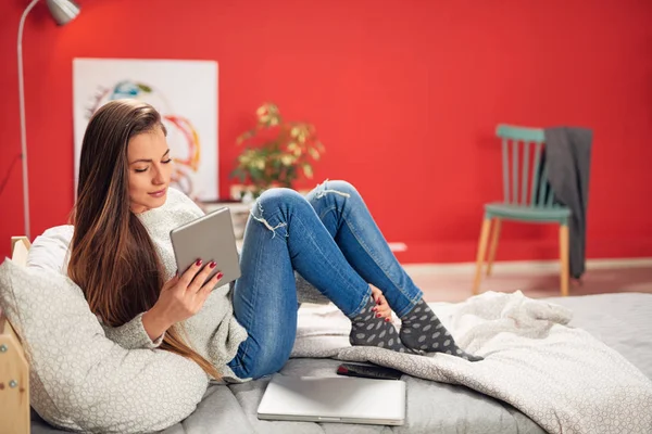 Wunderschöne lächelnde kaukasische Brünette lässig gekleidet im Schlafzimmer auf dem Bett sitzend und mit Tablet. — Stockfoto