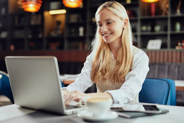 Bella bionda caucasica sorridente blogger donna vestita elegante casual seduta in mensa e utilizzando il computer portatile . — Foto Stock