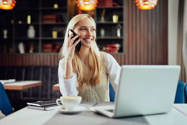 Jovem mulher de negócios loira caucasiana alegre vestida inteligente casual sentado no café e usando telefone inteligente . — Fotografia de Stock