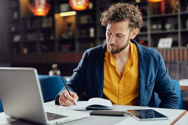 Unga allvarliga skäggiga kaukasiska bloggare klädd smart casual skriva anteckningar i agenda medan du sitter i cafeteria. — Stockfoto