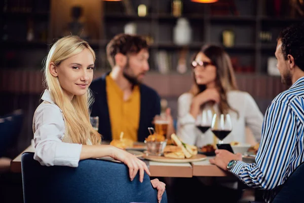 Jovem sorridente caucasiano mulher bonita loira vestida inteligente casual sentado à mesa no restaurante e olhando por cima do ombro. No fundo, seus amigos estão jantando . — Fotografia de Stock