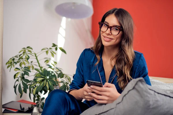 Charmante kaukasische Brünette in blauem Pyjama und mit Brille, die in die Kamera schaut, während sie im Schlafzimmer auf dem Bett sitzt und das Smartphone benutzt. Morgenstunde. — Stockfoto