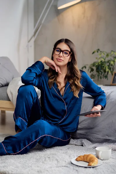 Beautiful Caucasian brunette in blue pajamas and with eyeglasses using tablet while sitting on the floor in bedroom. On floor is breeakfast. Morning time. — Stock Photo, Image