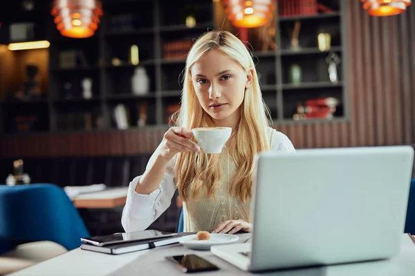 Mulher de negócios loira encantadora vestiu café casual inteligente bebendo no café e olhando para a câmera. Na mesa estão laptop, agenda e telefone inteligente . — Fotografia de Stock