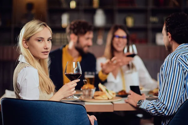 Portret van mooie blanke blonde vrouw zittend in restaurant en het houden van glas rode wijn. In de achtergrond vrienden het diner. — Stockfoto