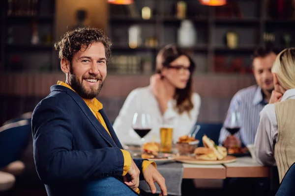 Jonge glimlachende man gekleed slim casual zitten in restaurant en kijken over de schouder. Op de achtergrond zijn vrienden aan het eten. — Stockfoto