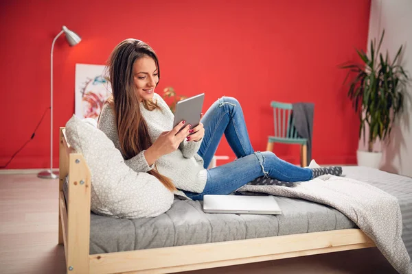 Hermosa morena caucásica sonriente vestida casual sentada en el dormitorio en la cama y usando la tableta . — Foto de Stock