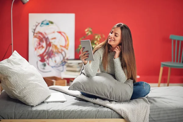 Hermosa morena caucásica sonriente vestida casual sentada en el dormitorio en la cama y usando la tableta . — Foto de Stock