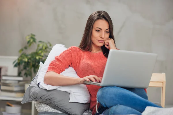 Beautiful Caucasian brunette sitting on the bed in bedroom and using laptop. — Stock Photo, Image