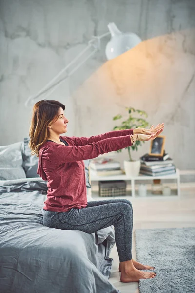 Encantadora morena caucásica sentada en la cama en el dormitorio y haciendo ejercicios de yoga . — Foto de Stock