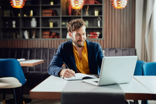 Joven blogger caucásico barbudo sonriente vestido elegante escritura casual notas en la agenda y mirando a la computadora portátil mientras está sentado en la cafetería . — Foto de Stock