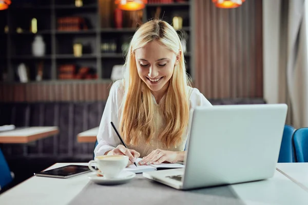 Charmante Kaukasische blonde zakenvrouw schrijven notities in notebook. Cafetaria interieur. — Stockfoto