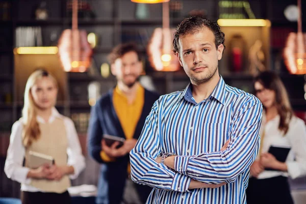 Mladý atraktivní arabský obchodník stojící v restauraci se zkříženýma rukama. V pozadí jeho úspěšný tým pózuje. — Stock fotografie