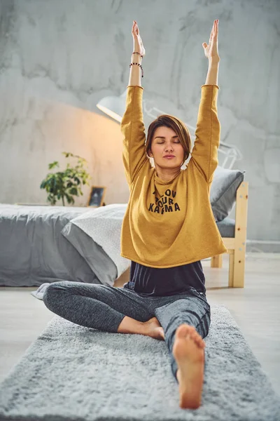 Hermosa morena caucásica sentada en la alfombra en el dormitorio y haciendo ejercicios de yoga . — Foto de Stock