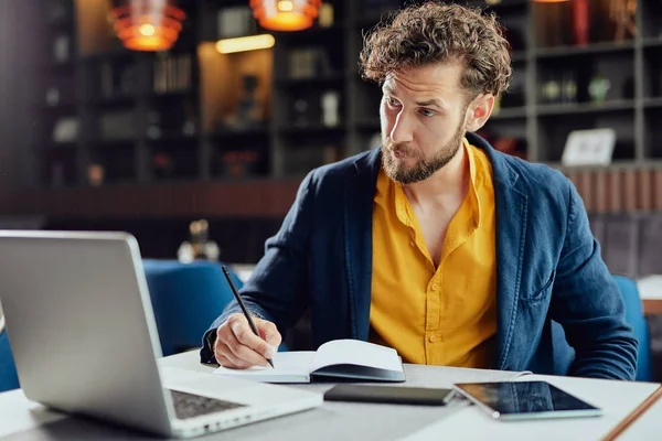 Young serious bearded Caucasian blogger dressed smart casual writing notes in agenda and looking at laptop while sitting in cafeteria. — Stockfoto