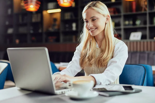 Bella bionda caucasica sorridente blogger donna vestita elegante casual seduta in mensa e utilizzando il computer portatile . — Foto Stock