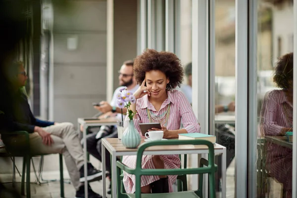 Smiling grogeous mixed race woman in pink striped dress sitting in cafe and using tablet. On table are cup of coffee, book and lemonade. — Stock Photo, Image