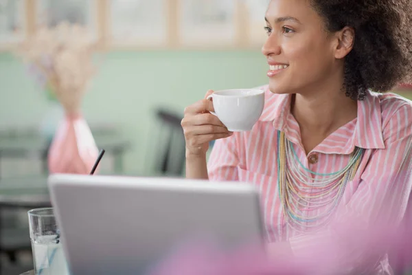 Charmante mixed race vrouw in gestreepte roze jurk aitting in Coffee Shop, koffie drinken en het gebruik van laptop. — Stockfoto