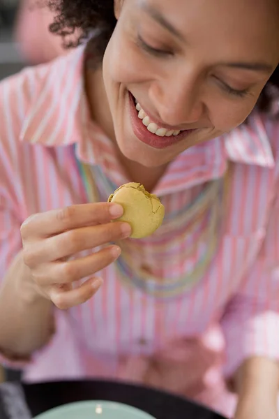 Primer plano de hermosa mujer de raza mixta sonriente en vestido a rayas rosa sentado en confitería y comer galleta . — Foto de Stock
