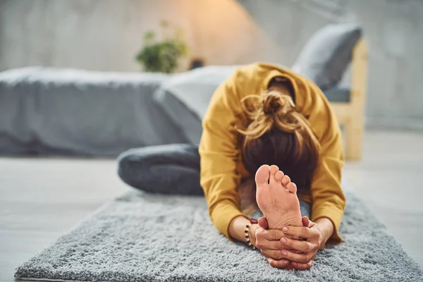 Caucásico morena estiramiento de la pierna mientras está sentado en la alfombra en el dormitorio . — Foto de Stock