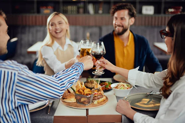 Four happy muliethnic freinds dressed smart casual cheering with alcohol while sitting at restaurant. — Stock Photo, Image