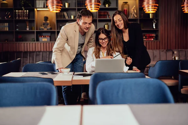 Charming buisnesswoman with brown hair and dressed elegant sitting at restaurant and looking at laptop. Next to her standing two colleagues and looking at laptop, too. — Stock Photo, Image