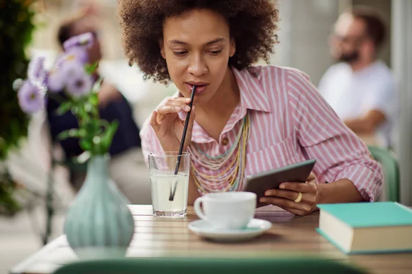 Prachtige mixed race vrouw in roze gestreepte jurk drinken limonade en het gebruik van Tablet zittend in café. — Stockfoto