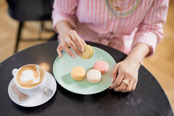 Vista superior de la mujer de raza mixta en vestido de rayas rosadas sosteniendo la galleta mientras está sentado en la pastelería. En la mesa hay café y plato con galletas . — Foto de Stock