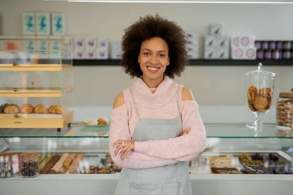 Belle femme souriante propriétaire mixte de pâtisserie vêtue de tablier debout avec les bras croisés dans la boutique . — Photo