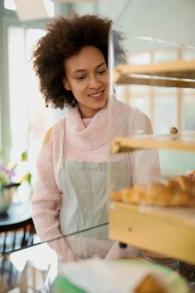 美丽的微笑混合种族女雇员在围裙摆姿势在糕点店nex计数器. — 图库照片