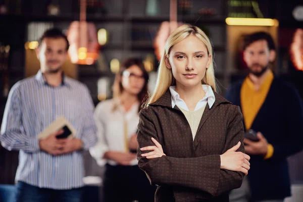 Portret pięknej kaukaski blond bizneswoman stoi w restauracji z ramionami skrzyżowane. W tle jej koledzy stojący i stwarzające. — Zdjęcie stockowe