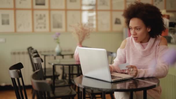 美丽的混血妇女在粉红色的高领毛衣使用笔记本电脑和信用卡在线购物 咖啡厅内饰 — 图库视频影像