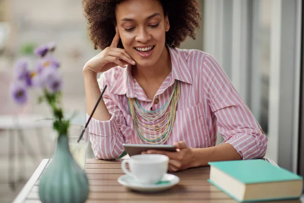 Sonriente hermosa mujer de raza mixta en vestido de rayas rosa sentado en la cafetería y el uso de la tableta. En la mesa hay una taza de café, libro y limonada. . — Foto de Stock
