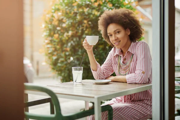 Mooie glimlachende mixed race vrouw in roze gestreepte jurk zitten in Cafe en enjoyingh haar koffie. — Stockfoto