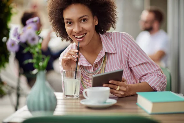 Prachtige mixed race vrouw in roze gestreepte jurk drinken limonade en het gebruik van Tablet zittend in café. — Stockfoto