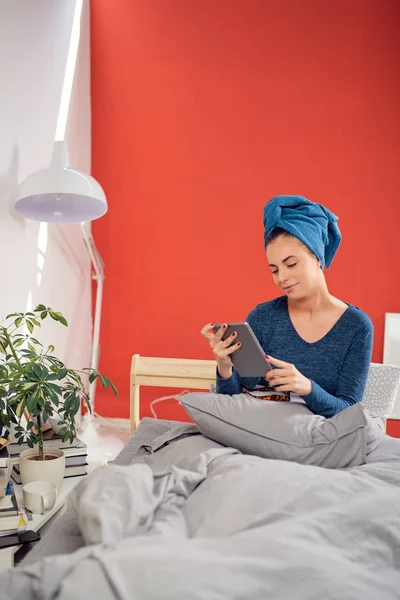 Hermosa morena con toalla en la cabeza usando tableta mientras está sentada en la cama en el dormitorio . — Foto de Stock