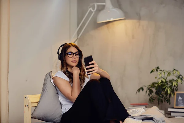 Bonita morena caucásica en pijama y con auriculares en la cabeza usando un teléfono inteligente tarde en la noche mientras está sentada en la cama en el dormitorio . — Foto de Stock