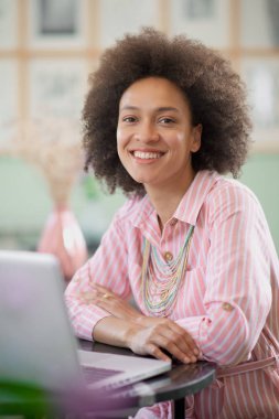 Kıvırcık saçlı güzel gülümseyen karışık yarış kadını, masanın üzerinde elleri ile kafede oturan ponk çizgili elbise giymiş. Masada dizüstü bilgisayar.